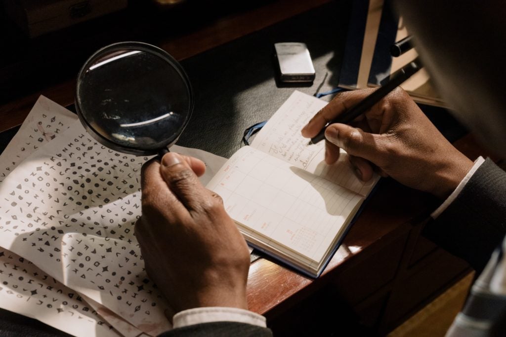 A picture of a person looking at a ciphertext with a magnifying glass and trying to decode the secret language -- for computer system, this process is known as encryption or cryptography and this article explores how it works, different types of encryption, use cases, and more.